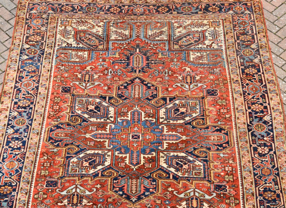A large antique Heriz room-size carpet displaying a prominent geometric central medallion in navy blue, terracotta red, and cream tones. View III