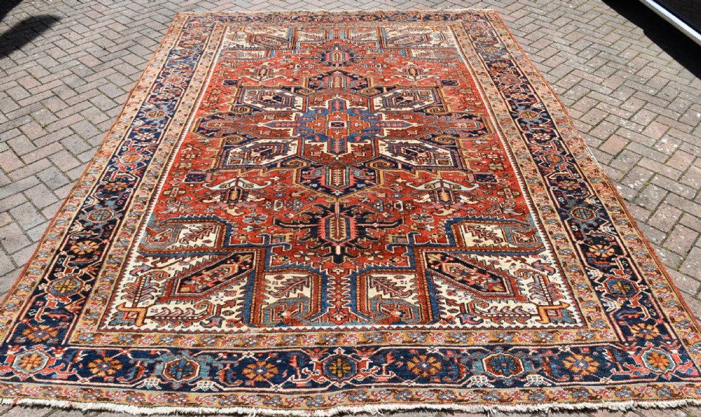 A large antique Heriz room-size carpet displaying a prominent geometric central medallion in navy blue, terracotta red, and cream tones. View IV