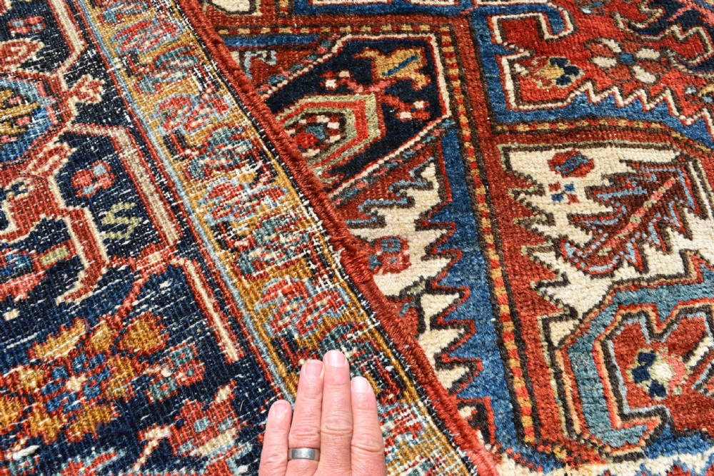 A large antique Heriz room-size carpet displaying a prominent geometric central medallion in navy blue, terracotta red, and cream tones. View V