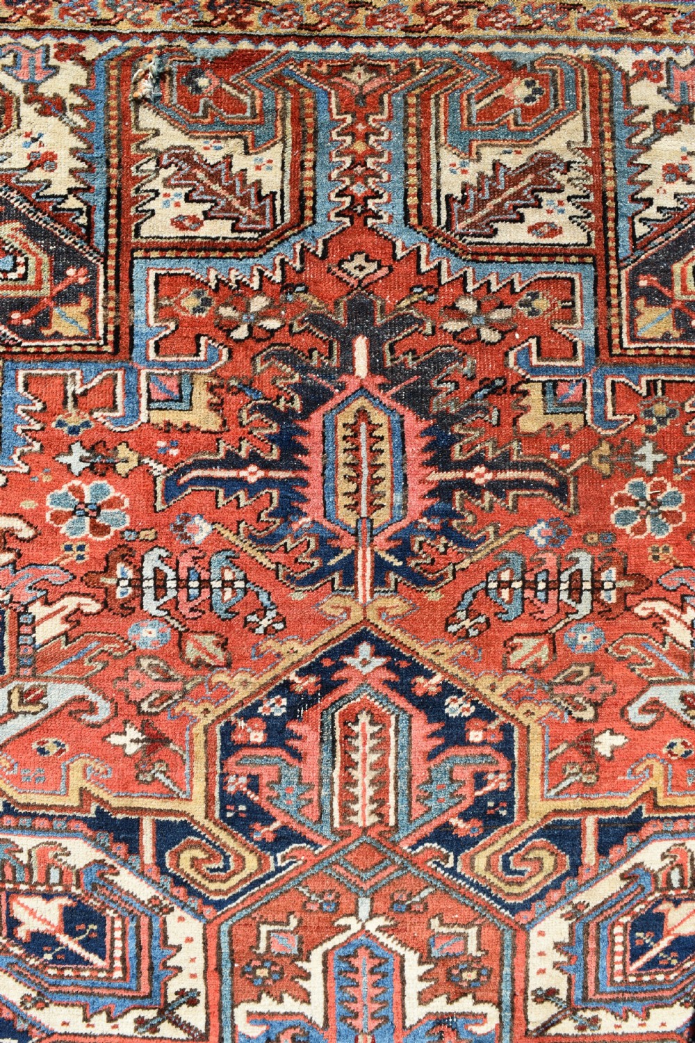 A large antique Heriz room-size carpet displaying a prominent geometric central medallion in navy blue, terracotta red, and cream tones. View VI