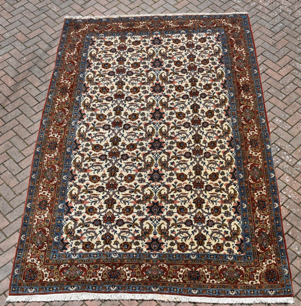 A large hand-knotted Persian Qum carpet featuring an all-over intricate floral vine motif on an ivory field, framed by multi-tiered red and blue decorative borders with fringe ends.