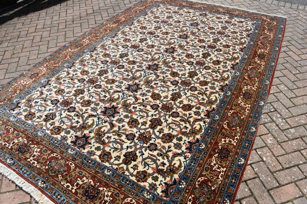 A large hand-knotted Persian Qum carpet featuring an all-over intricate floral vine motif on an ivory field, framed by multi-tiered red and blue decorative borders with fringe ends. View IV