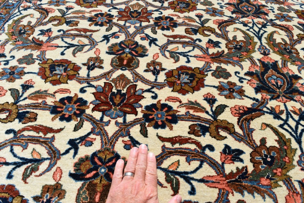A large hand-knotted Persian Qum carpet featuring an all-over intricate floral vine motif on an ivory field, framed by multi-tiered red and blue decorative borders with fringe ends. View V