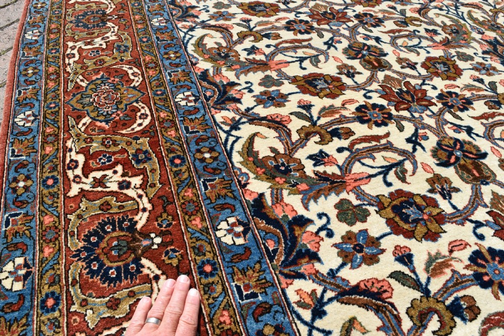 A large hand-knotted Persian Qum carpet featuring an all-over intricate floral vine motif on an ivory field, framed by multi-tiered red and blue decorative borders with fringe ends. View VII