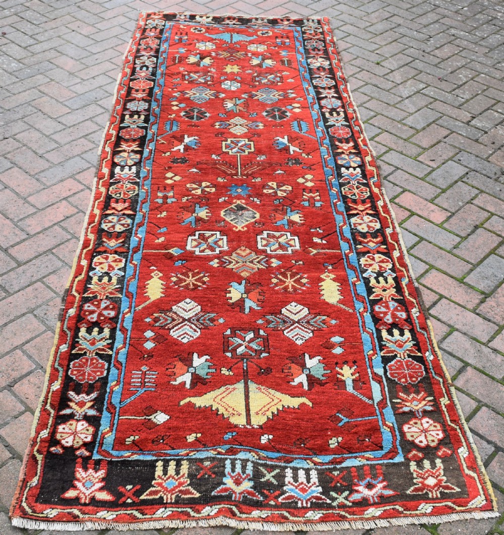 An antique long wool runner rug featuring a rich red field with geometric tribal motifs framed by a blue inner border and dark outer border.