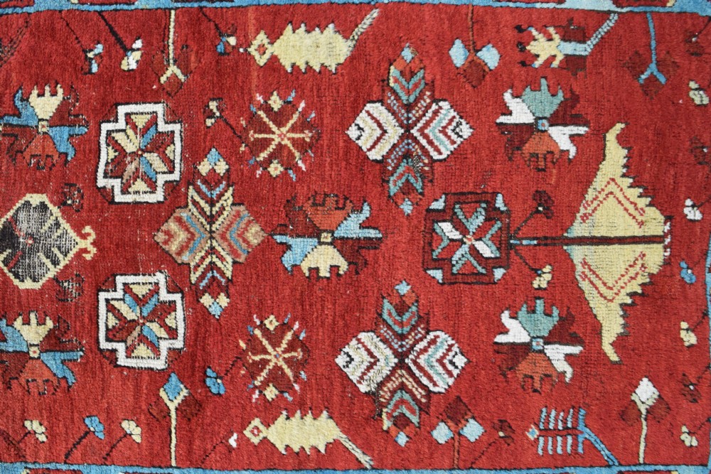 An antique long wool runner rug featuring a rich red field with geometric tribal motifs framed by a blue inner border and dark outer border. View III