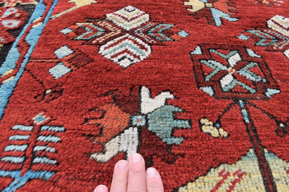 An antique long wool runner rug featuring a rich red field with geometric tribal motifs framed by a blue inner border and dark outer border. View VI
