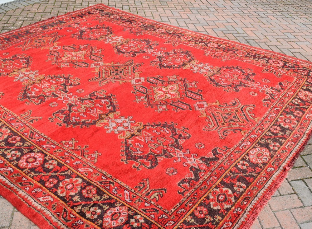 An antique hand-knotted Turkish Ushak wool carpet featuring a red field adorned with central geometric medallions and a patterned border. View IV