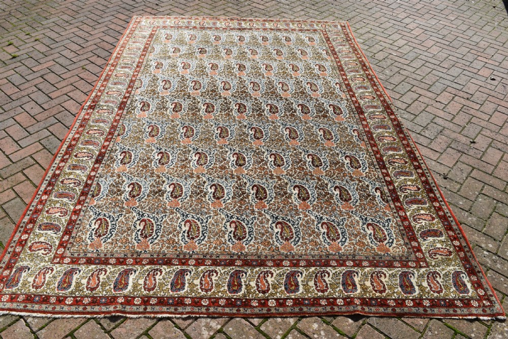 An antique hand-knotted Persian Qum carpet featuring an all-over repeating boteh paisley motif in cream, terracotta red, navy blue, and sage tones with a patterned border.