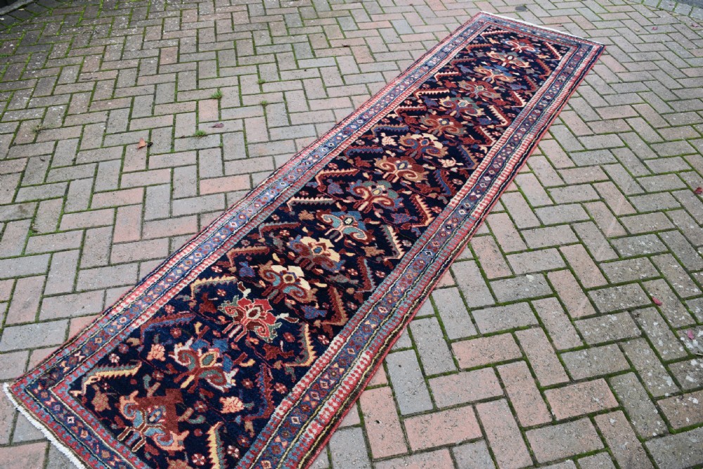 A long, narrow antique hand-knotted wool runner featuring intricate geometric floral motifs on a dark navy field bordered in terracotta and light blue. View III
