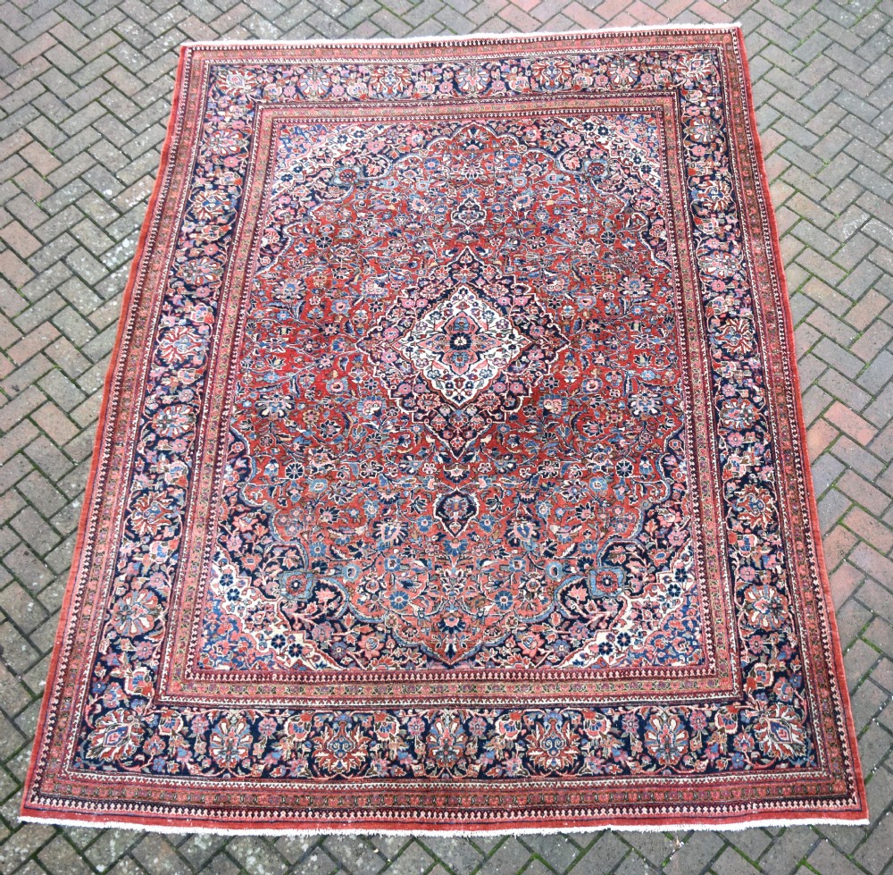 A large hand-knotted antique Kashan wool carpet featuring a central cream and blue medallion surrounded by dense floral patterns on a terracotta red field with decorative multi-tiered borders.