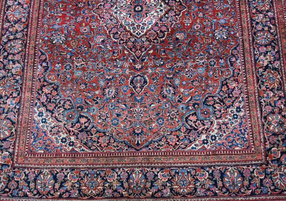 A large hand-knotted antique Kashan wool carpet featuring a central cream and blue medallion surrounded by dense floral patterns on a terracotta red field with decorative multi-tiered borders. View III