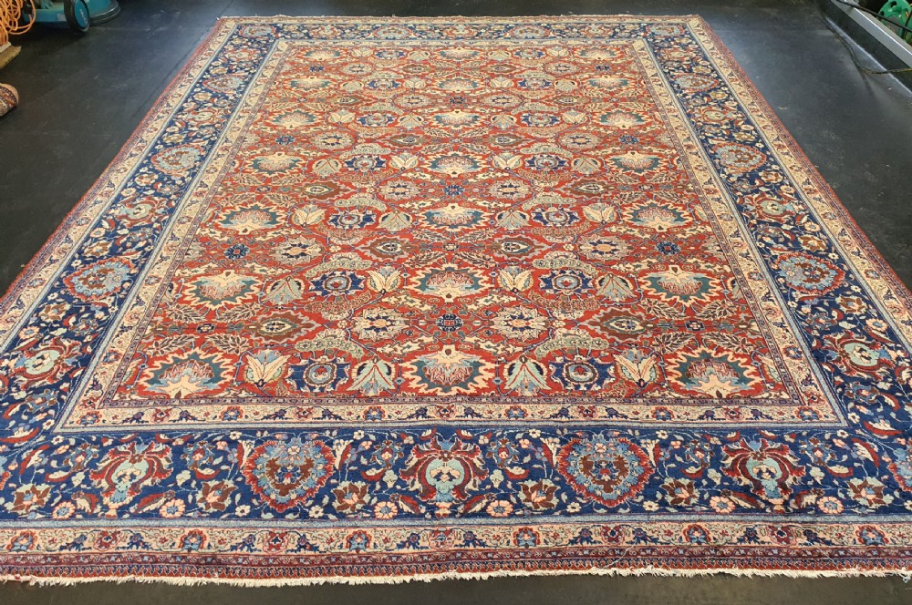 A large hand-knotted antique Tabriz carpet with an intricate floral motif across a red central field and a dark navy border.