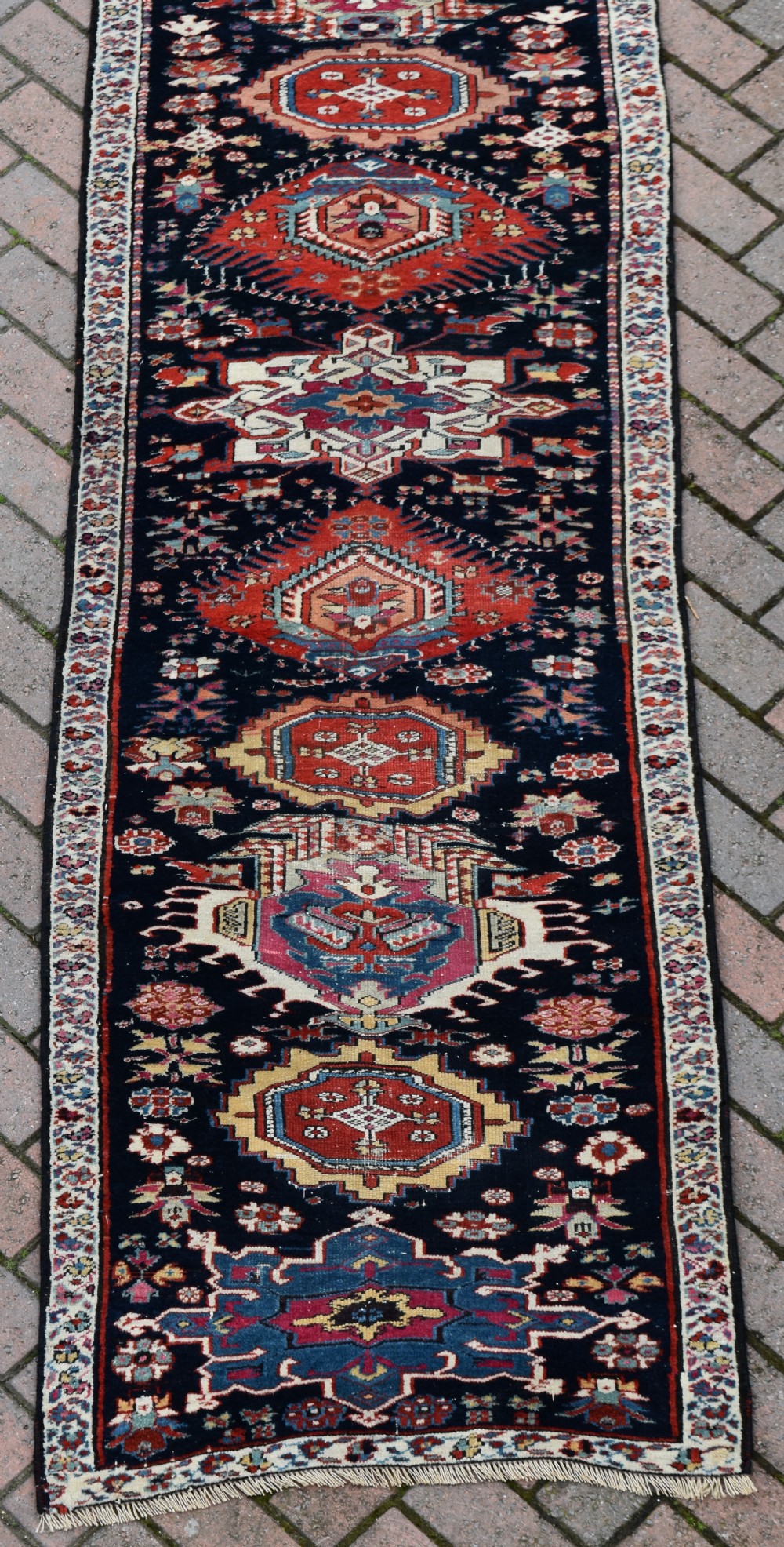 A long, narrow hand-knotted wool runner rug featuring repeating geometric medallions in red, blue, cream, and yellow set against a dark navy ground. View II