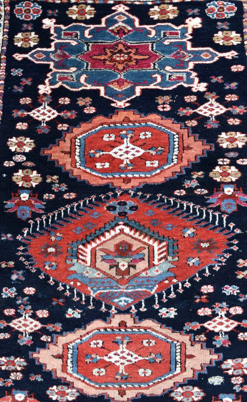 A long, narrow hand-knotted wool runner rug featuring repeating geometric medallions in red, blue, cream, and yellow set against a dark navy ground. View III