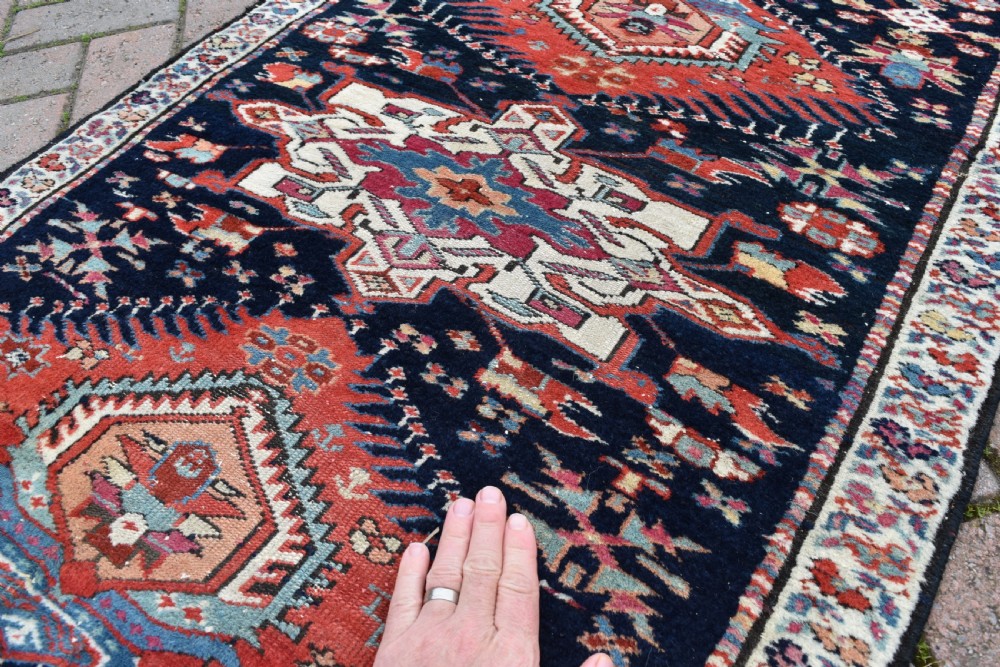 A long, narrow hand-knotted wool runner rug featuring repeating geometric medallions in red, blue, cream, and yellow set against a dark navy ground. View IV