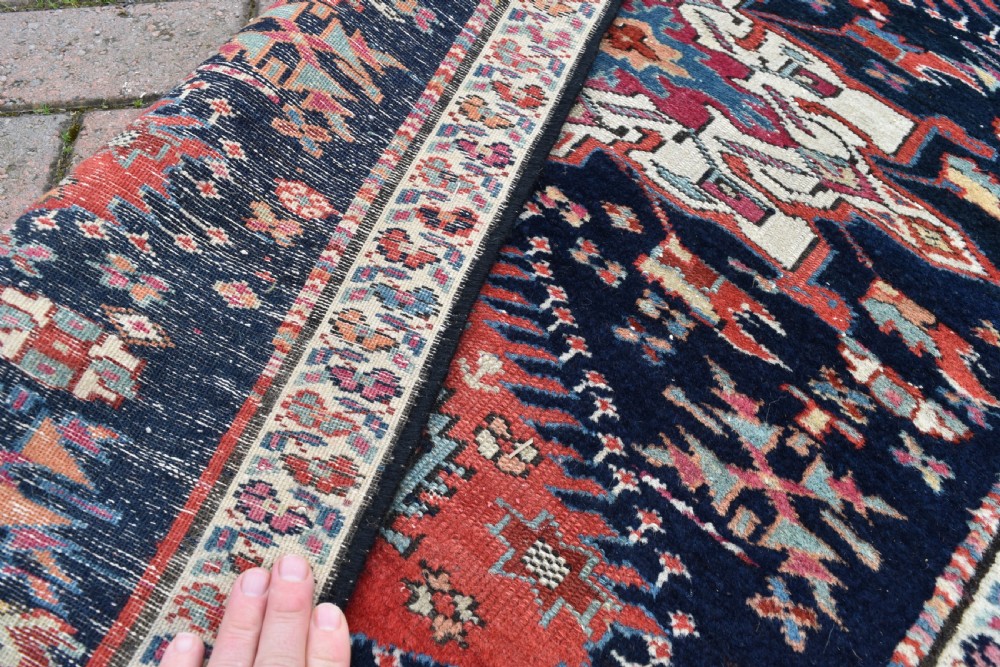 A long, narrow hand-knotted wool runner rug featuring repeating geometric medallions in red, blue, cream, and yellow set against a dark navy ground. View V