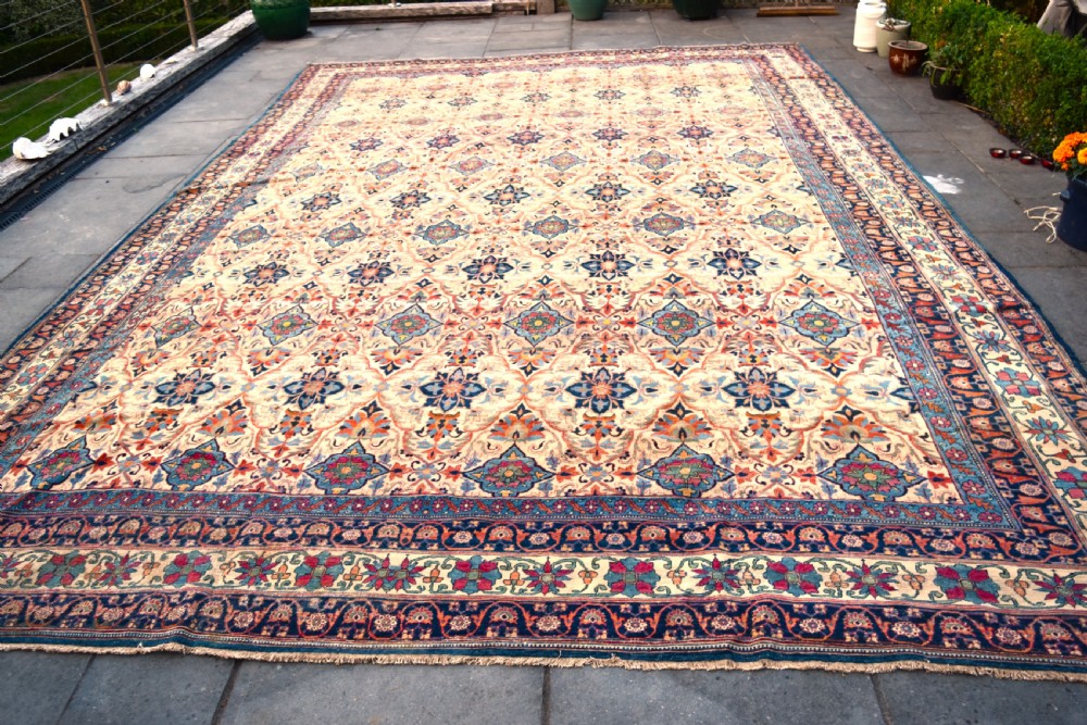 An oversized antique Kirman wool carpet featuring a cream field with intricate floral lattice motifs and a wide navy blue decorative border.