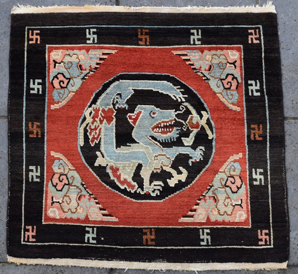 A square hand-knotted Tibetan wool rug featuring a central blue mythical beast inside a circular medallion on a red field with black geometric borders.