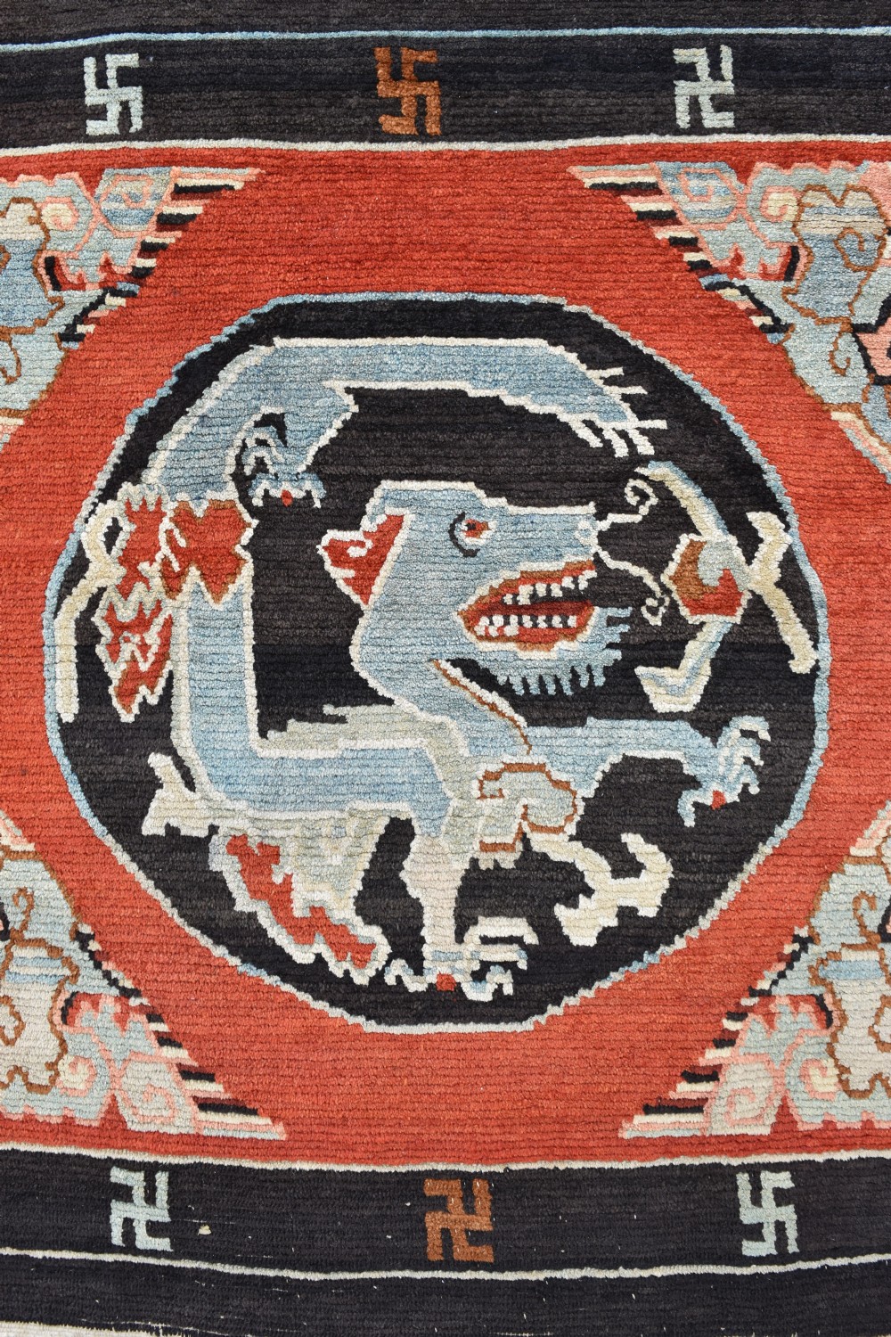 A square hand-knotted Tibetan wool rug featuring a central blue mythical beast inside a circular medallion on a red field with black geometric borders. View II