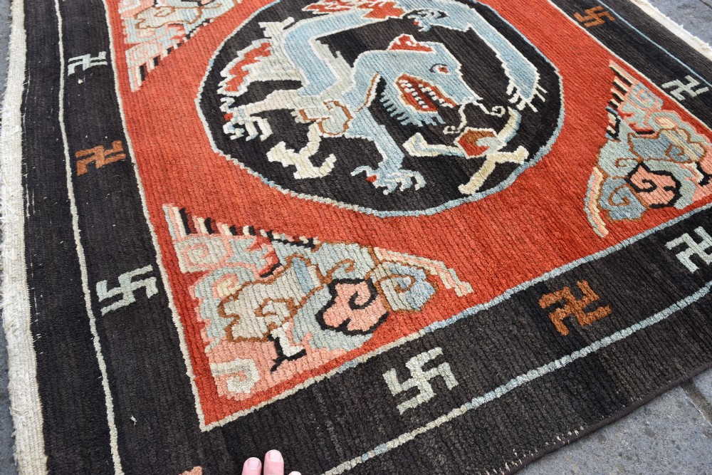 A square hand-knotted Tibetan wool rug featuring a central blue mythical beast inside a circular medallion on a red field with black geometric borders. View IV