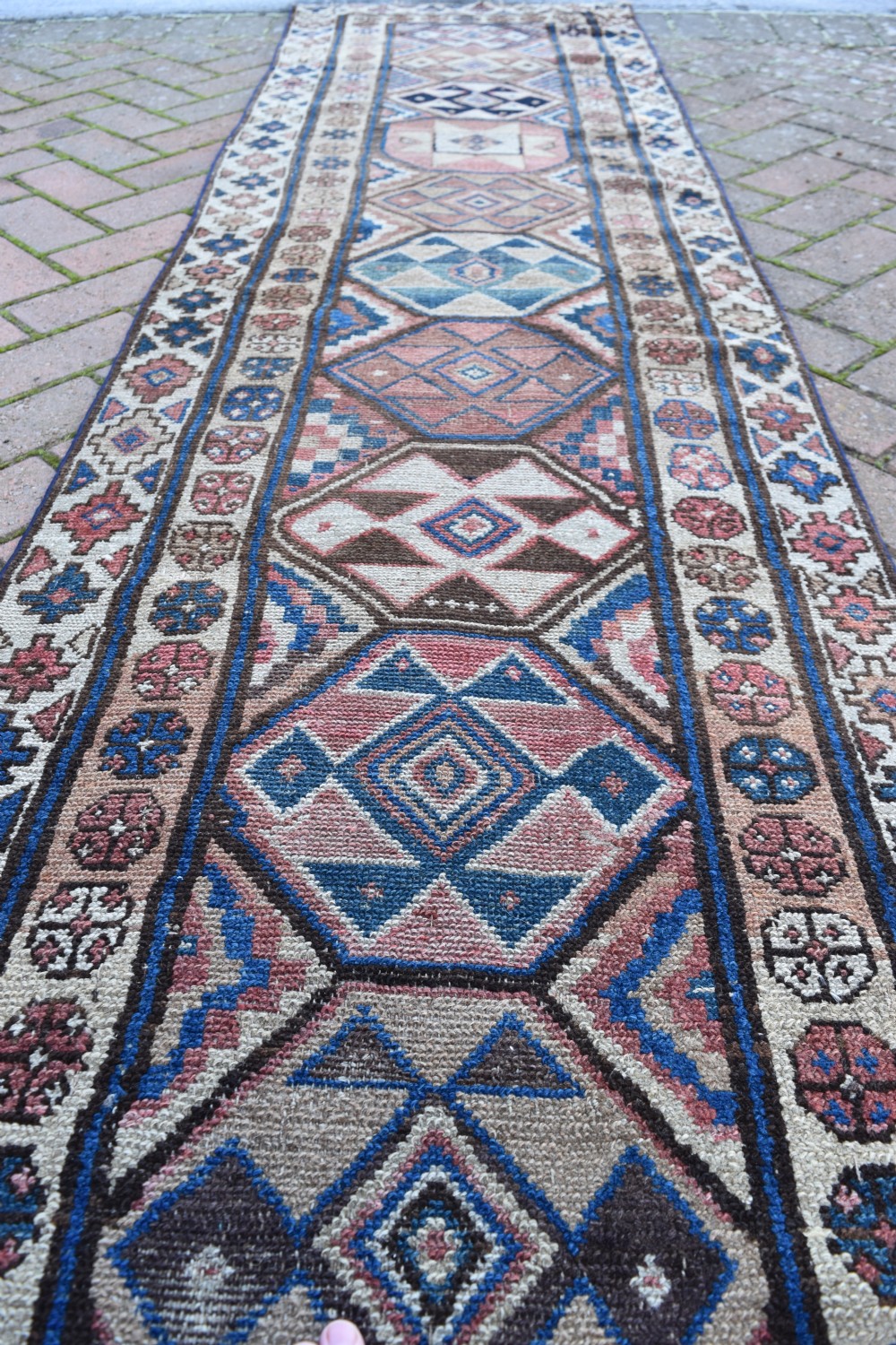 An exceptionally long, narrow hand-knotted wool runner rug featuring a series of geometric diamond medallions in red, navy blue, and cream along a decorated field with multi-banded patterned borders. View IX