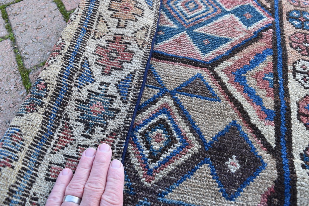 An exceptionally long, narrow hand-knotted wool runner rug featuring a series of geometric diamond medallions in red, navy blue, and cream along a decorated field with multi-banded patterned borders. View X