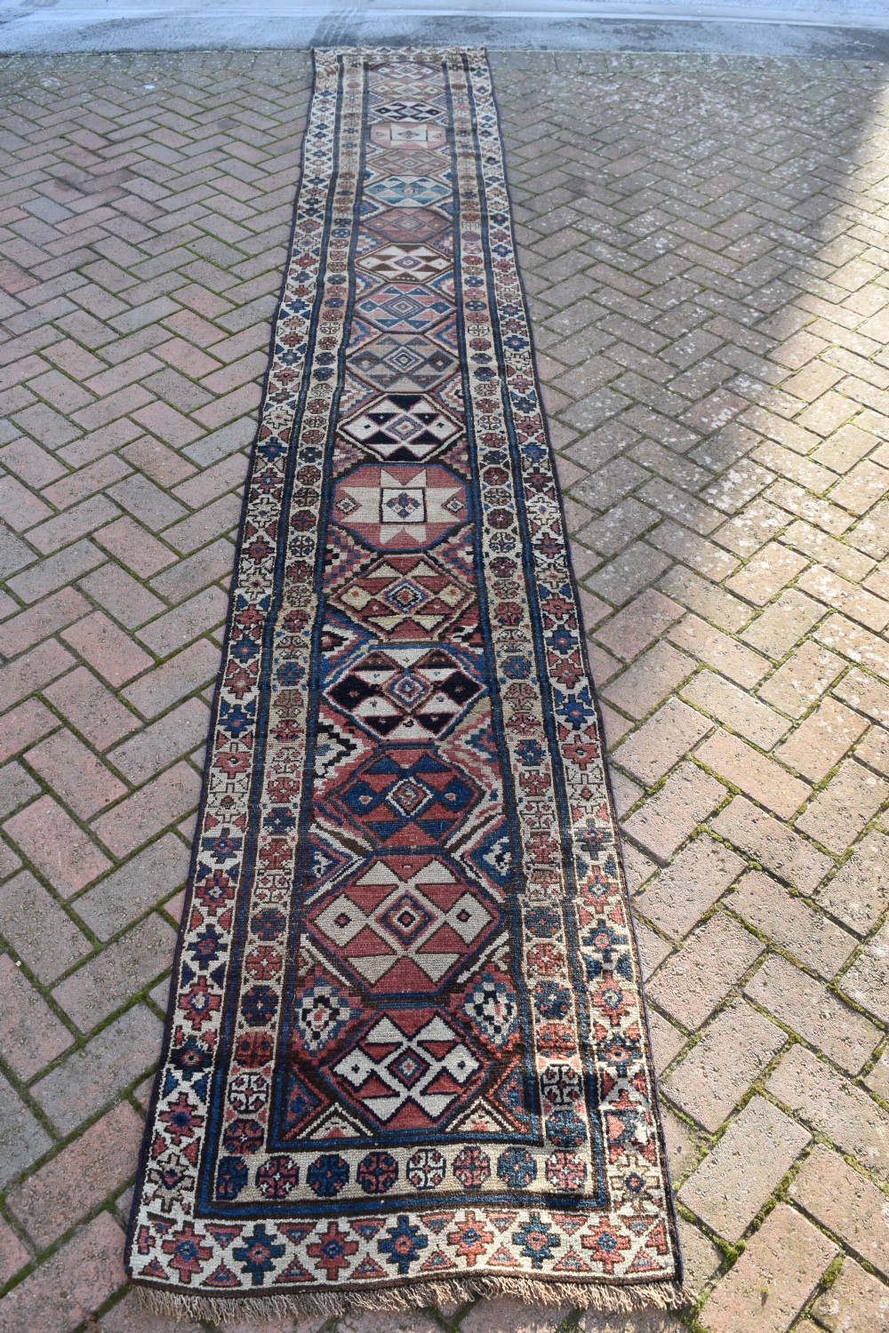 An exceptionally long, narrow hand-knotted wool runner rug featuring a series of geometric diamond medallions in red, navy blue, and cream along a decorated field with multi-banded patterned borders. View XI