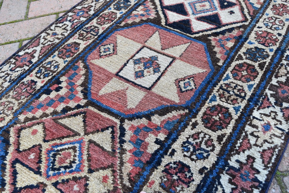 An exceptionally long, narrow hand-knotted wool runner rug featuring a series of geometric diamond medallions in red, navy blue, and cream along a decorated field with multi-banded patterned borders. View V