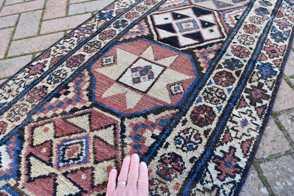 An exceptionally long, narrow hand-knotted wool runner rug featuring a series of geometric diamond medallions in red, navy blue, and cream along a decorated field with multi-banded patterned borders. View VII