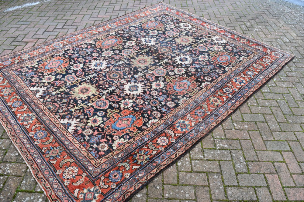A large rectangular hand-knotted wool carpet featuring a dark navy central field filled with geometric floral motifs and framed by a wide terracotta border. View III