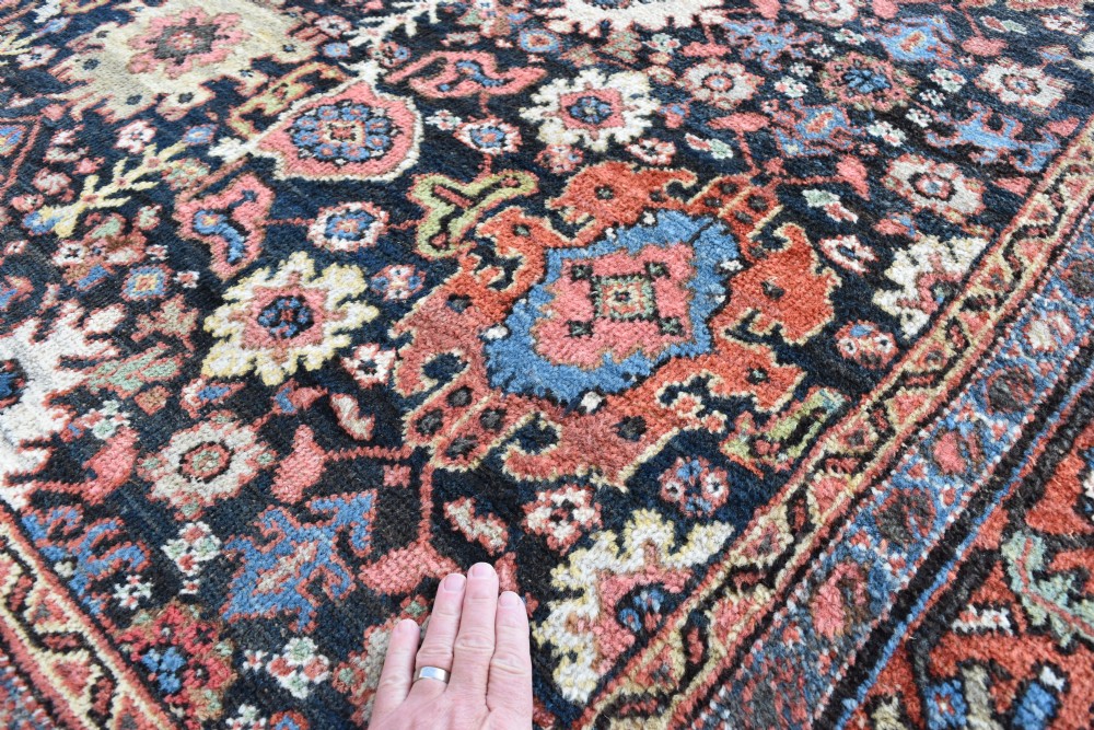 A large rectangular hand-knotted wool carpet featuring a dark navy central field filled with geometric floral motifs and framed by a wide terracotta border. View IV