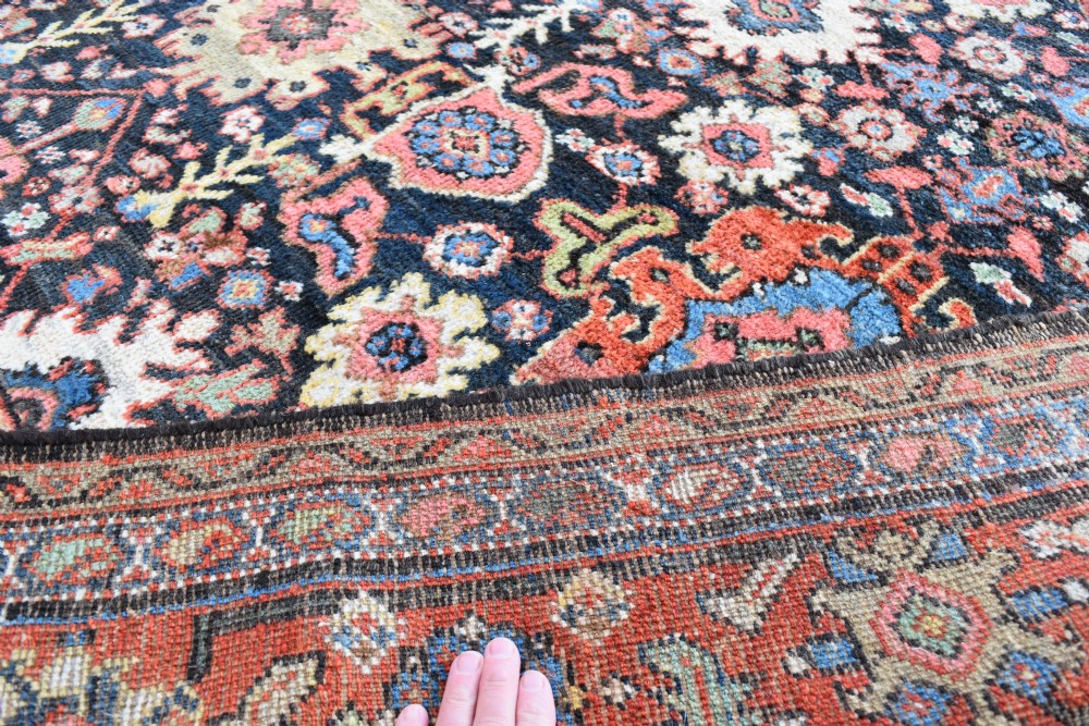 A large rectangular hand-knotted wool carpet featuring a dark navy central field filled with geometric floral motifs and framed by a wide terracotta border. View V