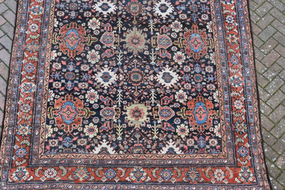 A large rectangular hand-knotted wool carpet featuring a dark navy central field filled with geometric floral motifs and framed by a wide terracotta border. View VI