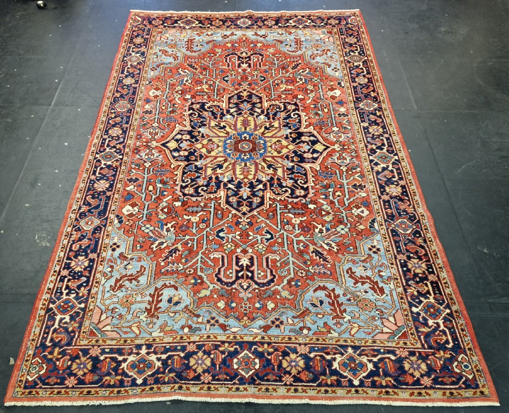 An antique hand-knotted Persian Heriz carpet with a central geometric navy medallion on a terracotta red field with light blue spandrels.