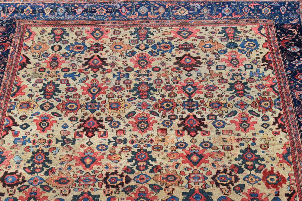 A large square hand-knotted antique Ferahan wool carpet featuring an all-over repeating geometric and floral pattern on a cream field, framed by a wide navy blue border. View IV