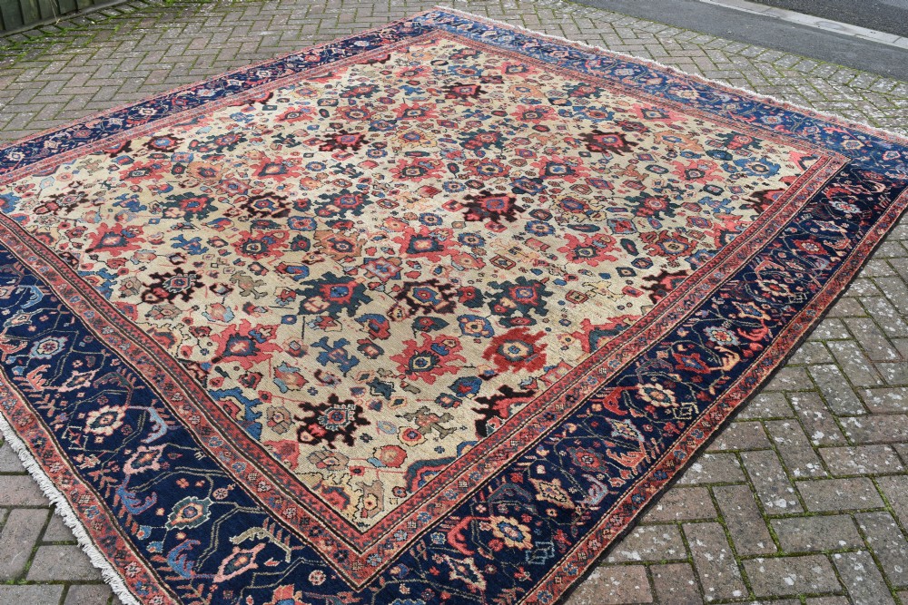 A large square hand-knotted antique Ferahan wool carpet featuring an all-over repeating geometric and floral pattern on a cream field, framed by a wide navy blue border. View V