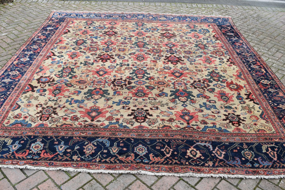 A large square hand-knotted antique Ferahan wool carpet featuring an all-over repeating geometric and floral pattern on a cream field, framed by a wide navy blue border. View VI