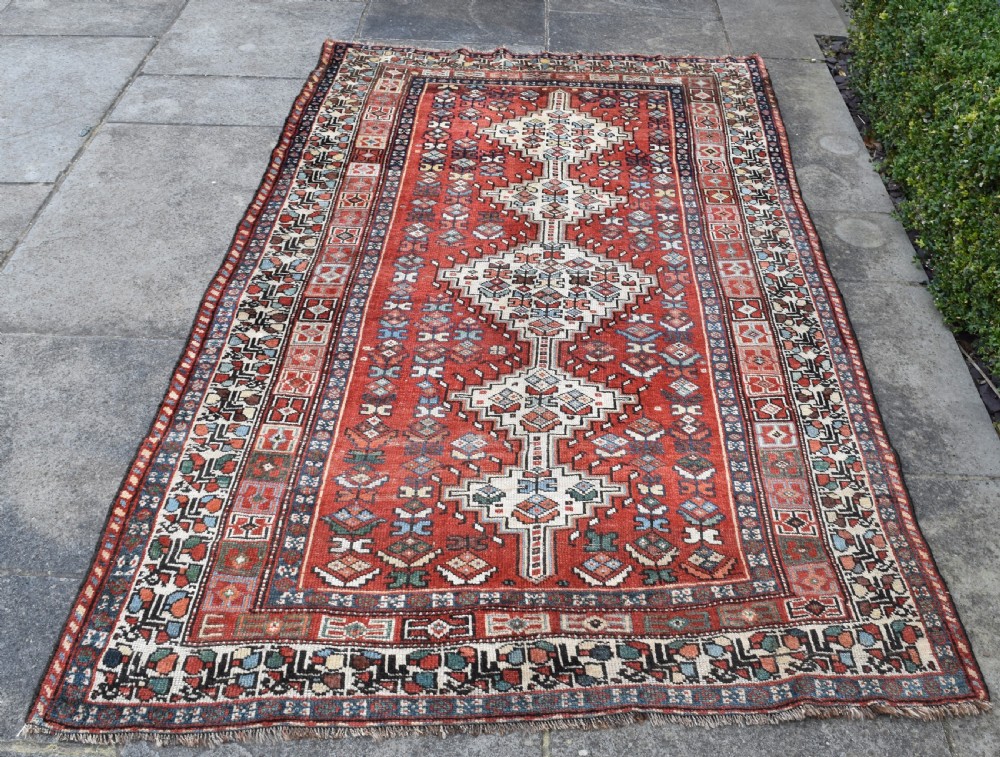 An antique hand-knotted Luri wool carpet showcasing a terracotta red field with central cream diamond medallions and geometric tribal borders. View II