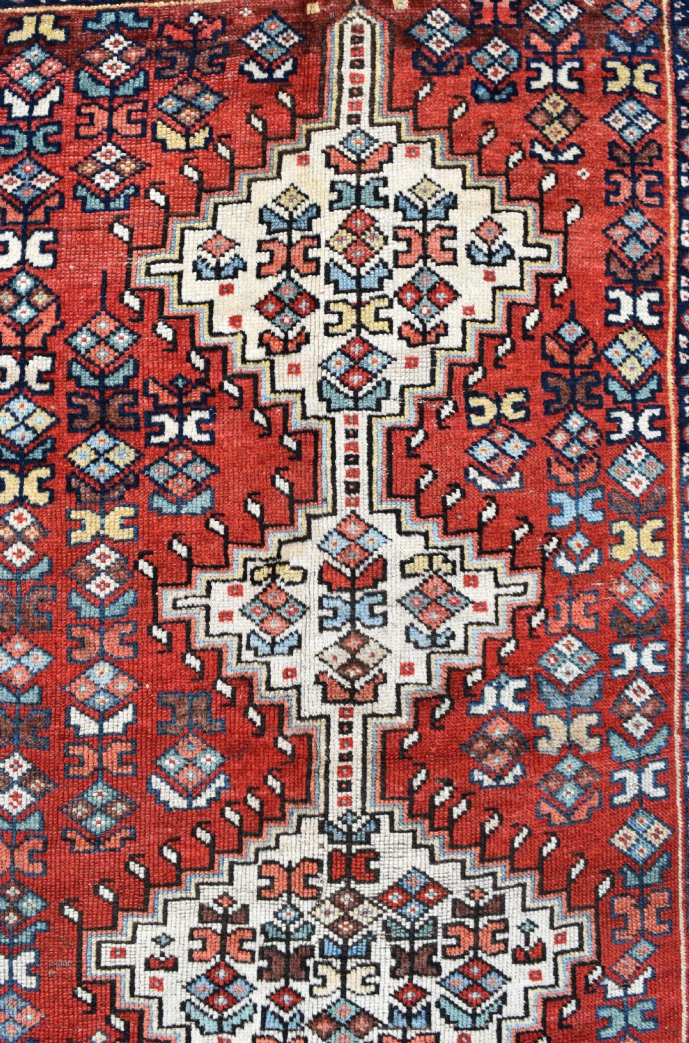 An antique hand-knotted Luri wool carpet showcasing a terracotta red field with central cream diamond medallions and geometric tribal borders. View III
