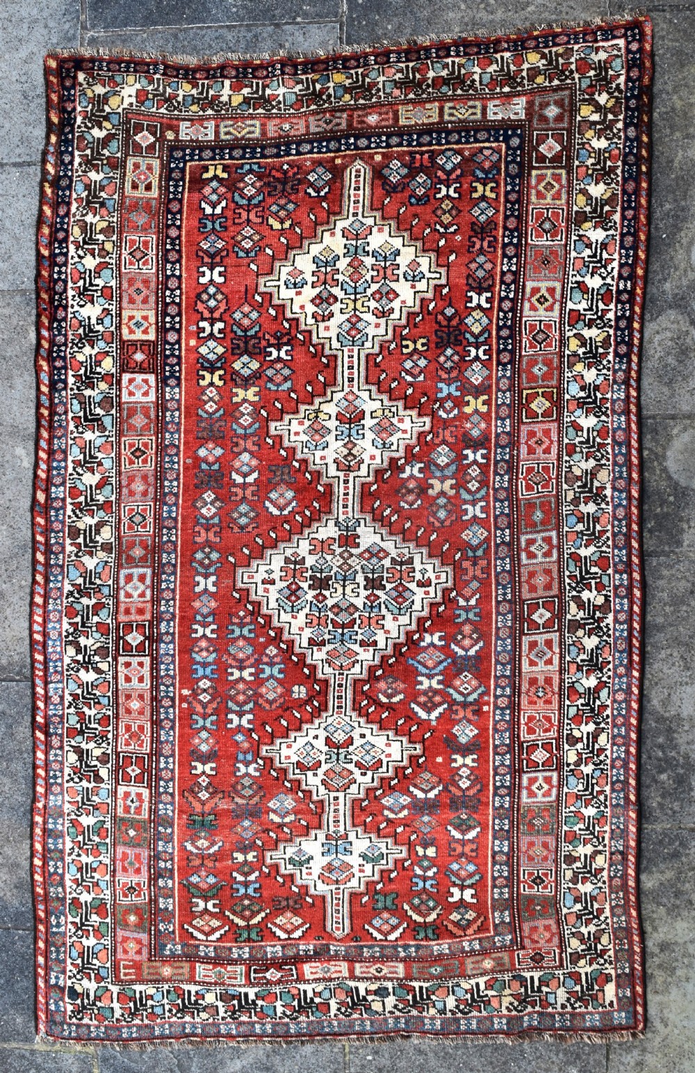 An antique hand-knotted Luri wool carpet showcasing a terracotta red field with central cream diamond medallions and geometric tribal borders. View IV
