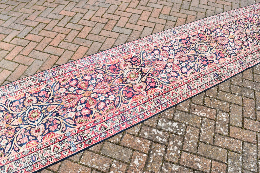 An exceptionally long and narrow hand-knotted antique Kirman rug runner featuring detailed floral medallion motifs in pink, navy blue, and cream. View V