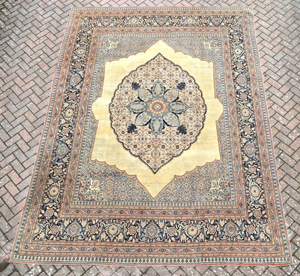 A large hand-knotted antique Persian Hajji Jalili carpet featuring a central star-shaped medallion on a cream field surrounded by dark blue floral borders.