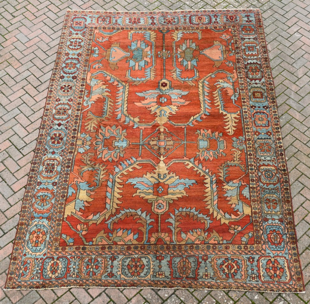 A large hand-knotted Heriz carpet featuring a rust red central field with geometric medallions in light blue, cream, and olive green, framed by a multi-tiered patterned border.