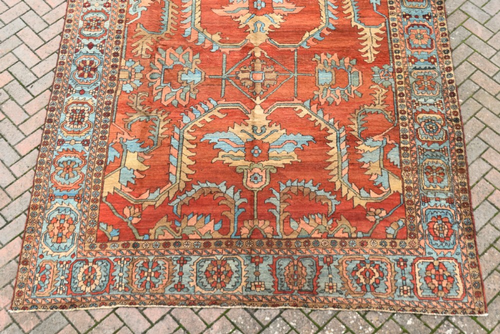A large hand-knotted Heriz carpet featuring a rust red central field with geometric medallions in light blue, cream, and olive green, framed by a multi-tiered patterned border. View II