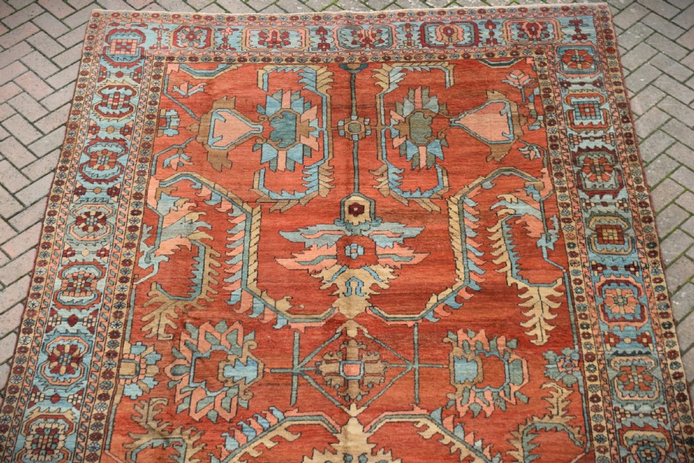 A large hand-knotted Heriz carpet featuring a rust red central field with geometric medallions in light blue, cream, and olive green, framed by a multi-tiered patterned border. View III
