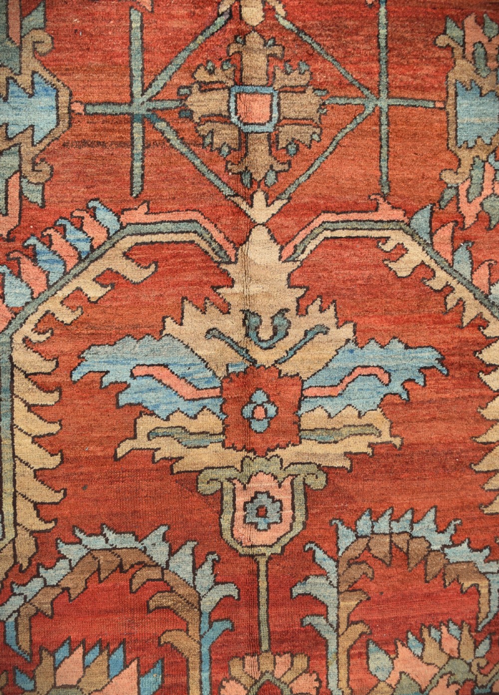 A large hand-knotted Heriz carpet featuring a rust red central field with geometric medallions in light blue, cream, and olive green, framed by a multi-tiered patterned border. View IV