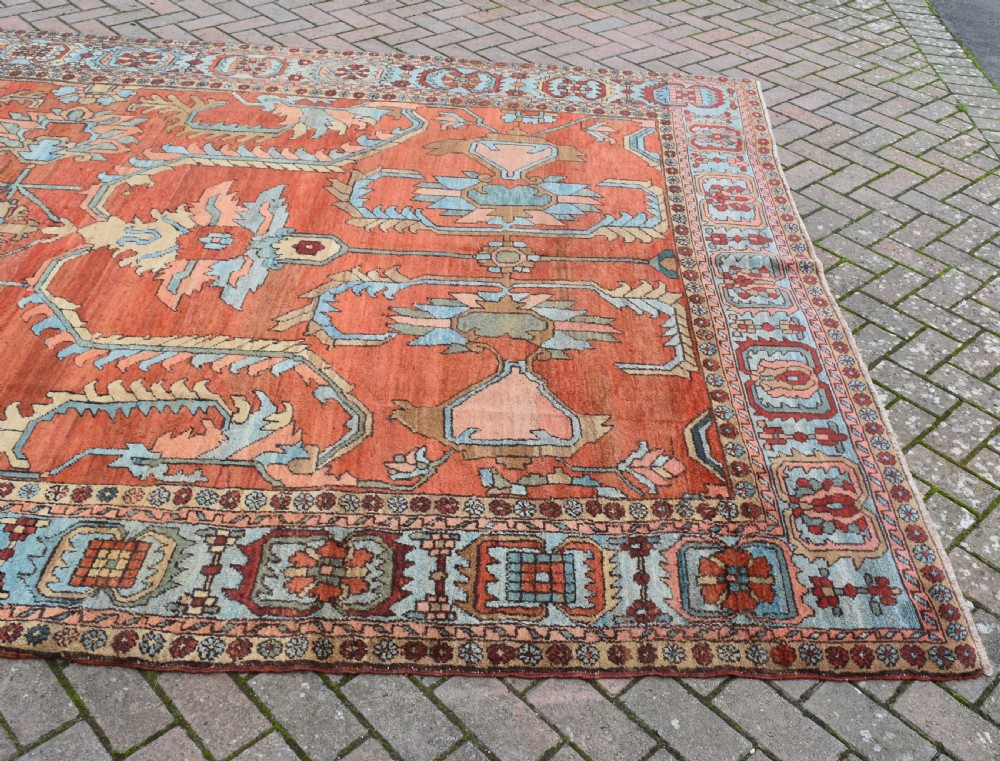 A large hand-knotted Heriz carpet featuring a rust red central field with geometric medallions in light blue, cream, and olive green, framed by a multi-tiered patterned border. View V