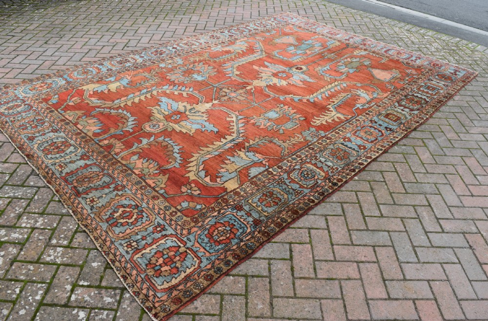A large hand-knotted Heriz carpet featuring a rust red central field with geometric medallions in light blue, cream, and olive green, framed by a multi-tiered patterned border. View VI