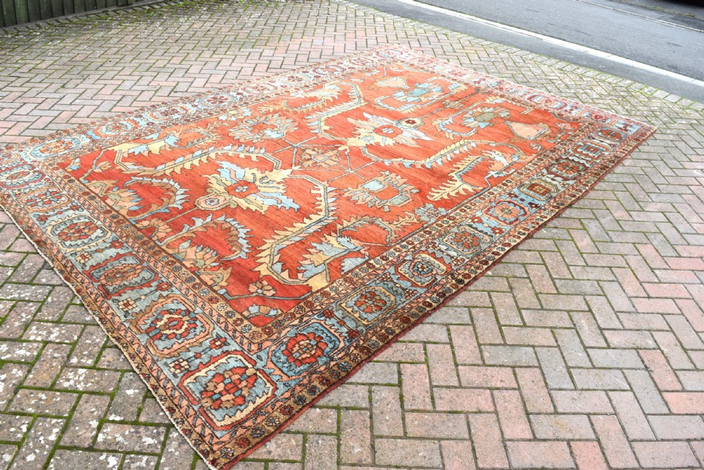A large hand-knotted Heriz carpet featuring a rust red central field with geometric medallions in light blue, cream, and olive green, framed by a multi-tiered patterned border. View VII